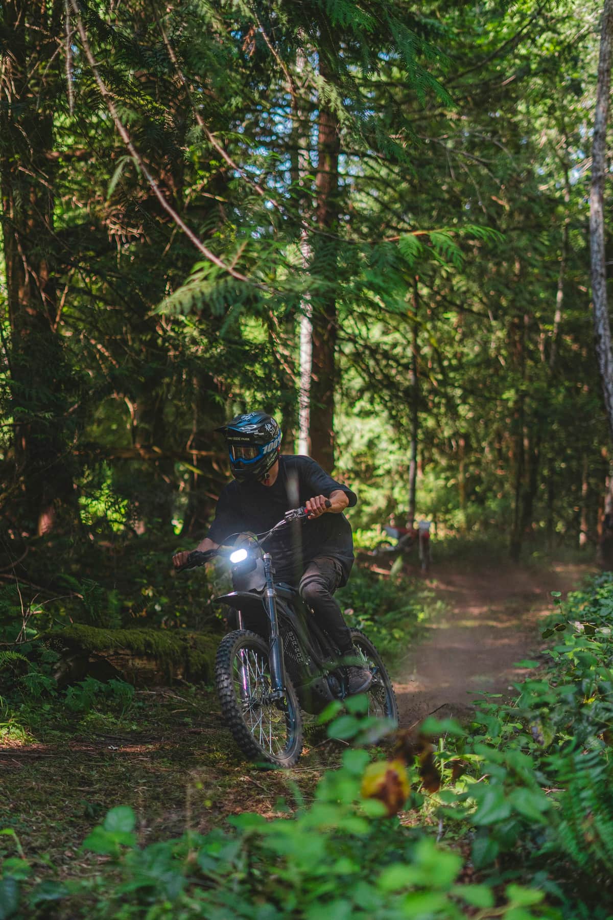 A rider on a dirt bike climbing a narrow trail through a dense forest.