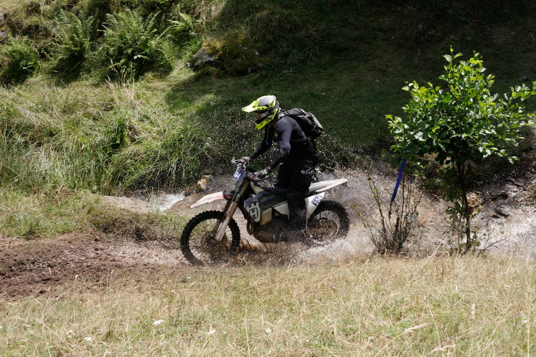 A rider on a dirt bike crossing a shallow creek on a grassy trail.